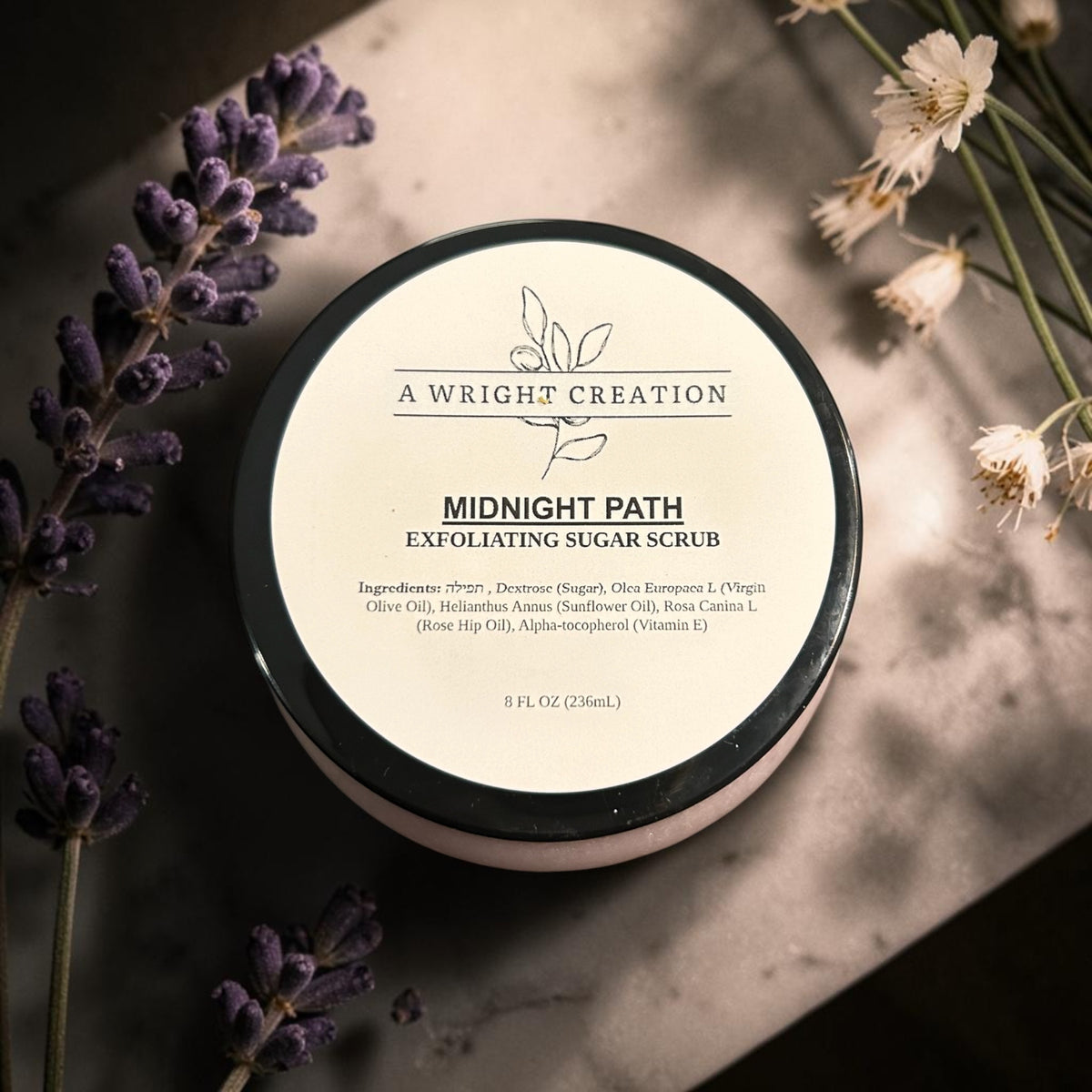 A round cosmetic jar of A Wright Creation Midnight Path Exfoliating Sugar Scrub, styled on a textured gray surface with dried white flowers and purple lavender sprigs.