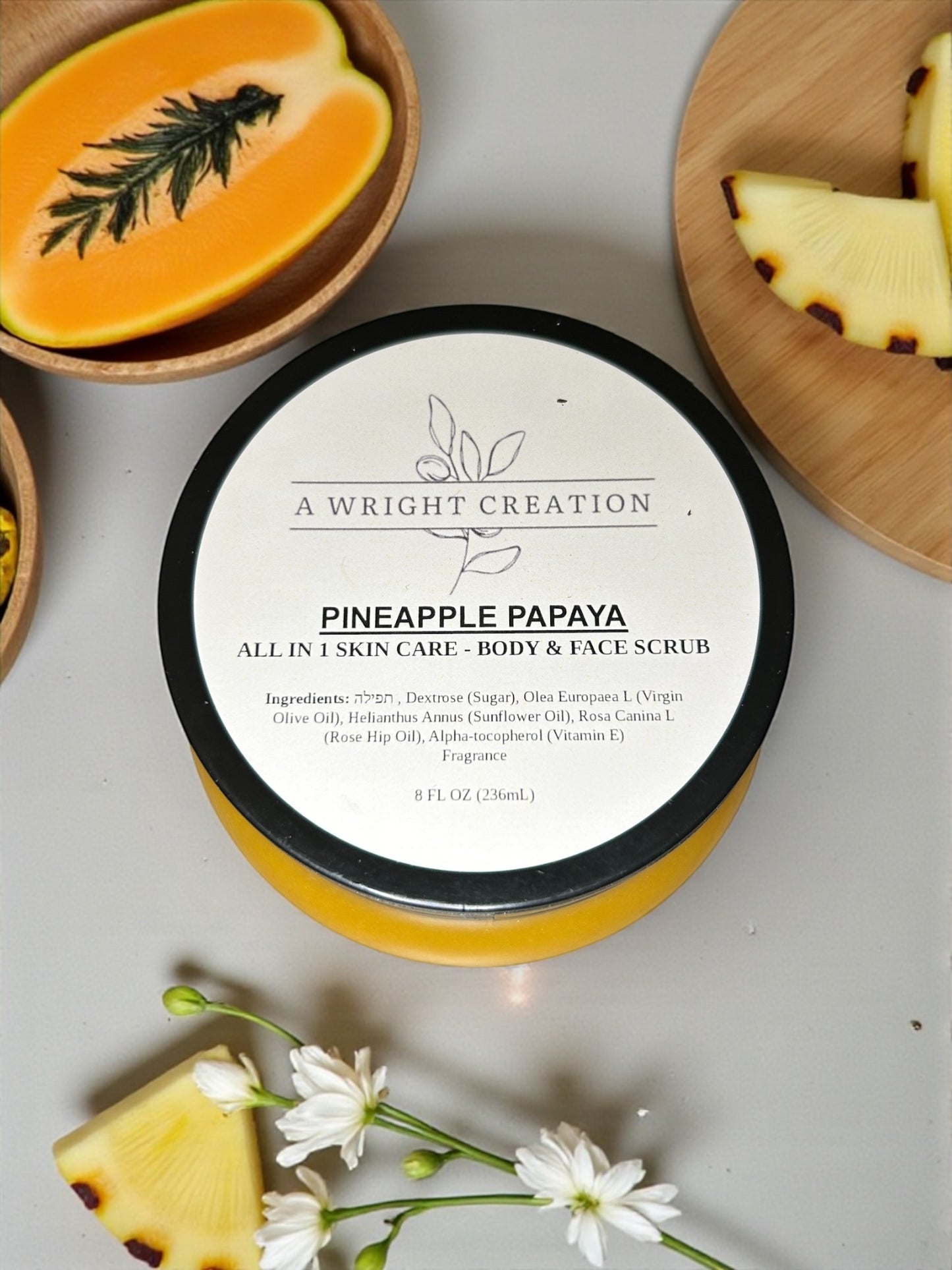 Pineapple Papaya skin care scrub container with ingredients listed, surrounded by pineapple slices and flowers on a light background.