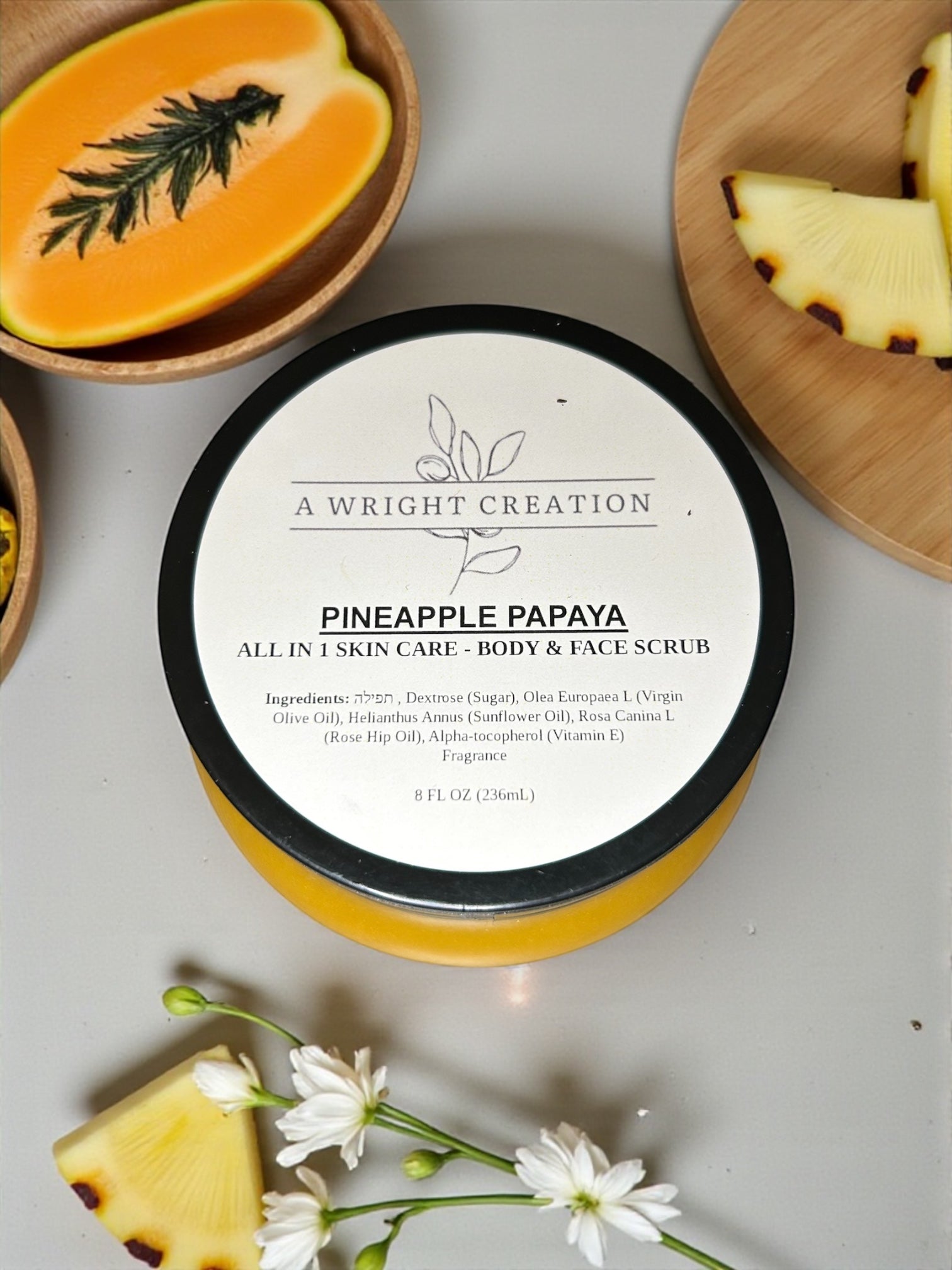 Pineapple Papaya skin care scrub container with ingredients listed, surrounded by pineapple slices and flowers on a light background.