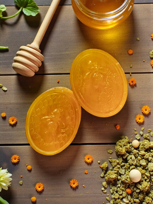 Two yellow honeycombs with a wooden honey dipper on a wooden surface with flowers and herbs.